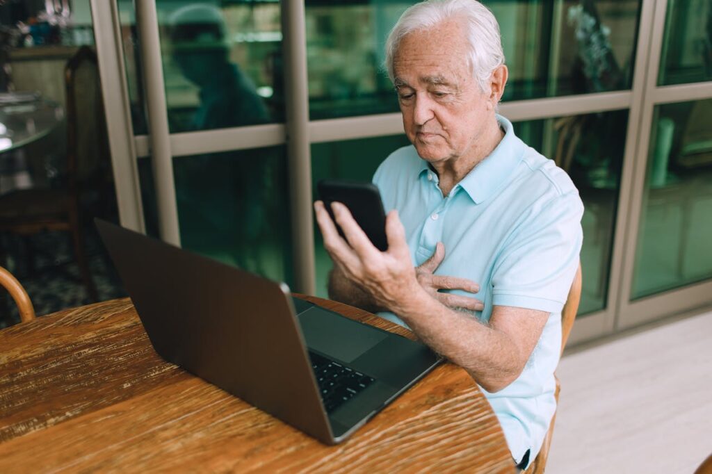 Elderly man sitting at a table using a smartphone and laptop, showcasing technology use.
